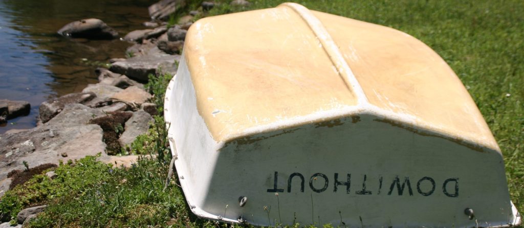 A white, plastic boat flipped upsidedown on the grassy shore of a rocky lake. The boat is inscribed with the words "DO WITHOUT." This image was taken at International Dai Bosatsu Zendo Kongo-ji in Livingston Manor, New York, USA.